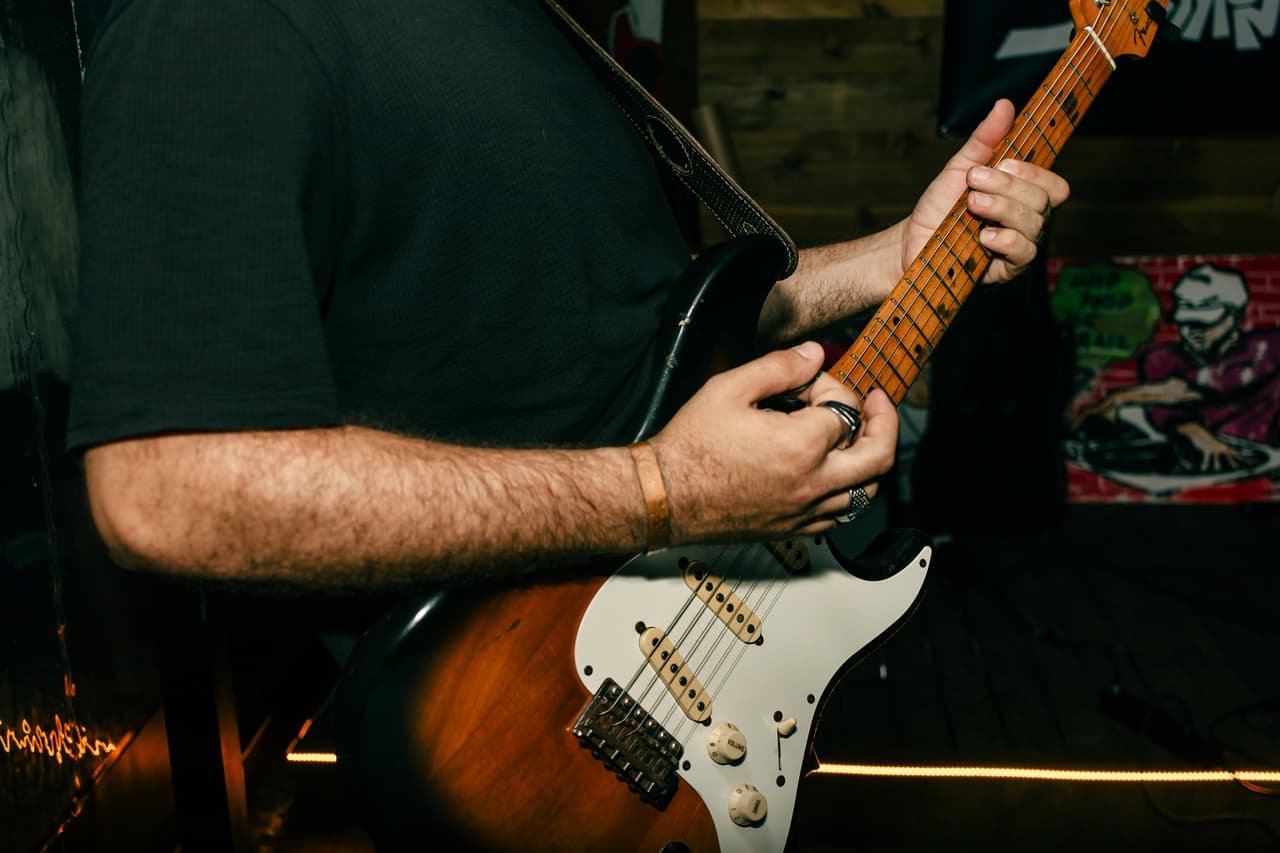Close das mãos de um guitarrista dedilhando uma guitarra de corpo sunburst e escudo branco