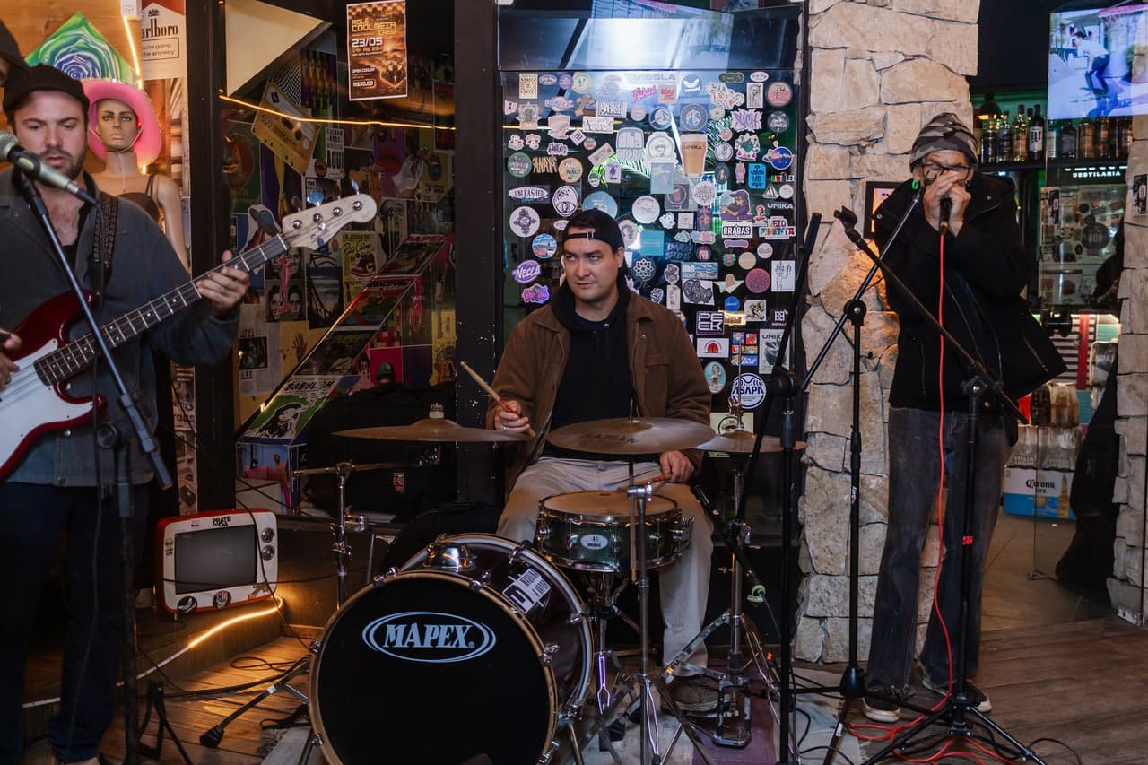 Banda no palco com guitarrista, baterista e vocalista diante de uma parede coberta de adesivos coloridos