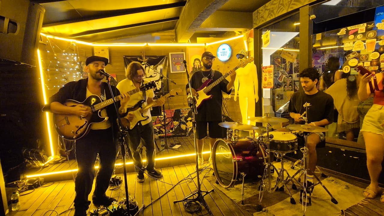 Banda completa com guitarras, baixo e bateria tocando num palco de madeira cercado por fita de luz amarela