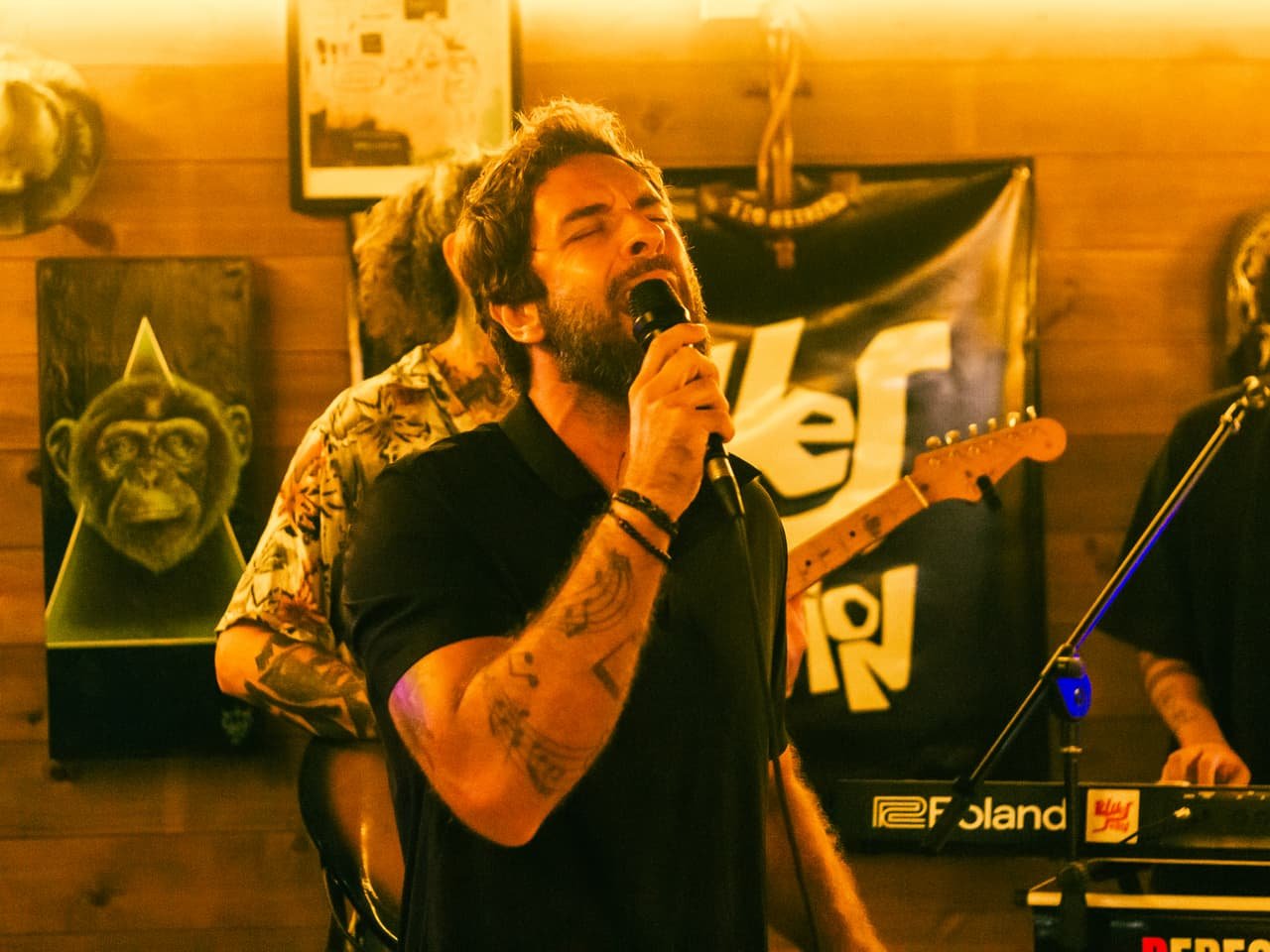 Cantor tatuado canta ao microfone na frente do banner Blues Session, com guitarrista e teclado ao lado