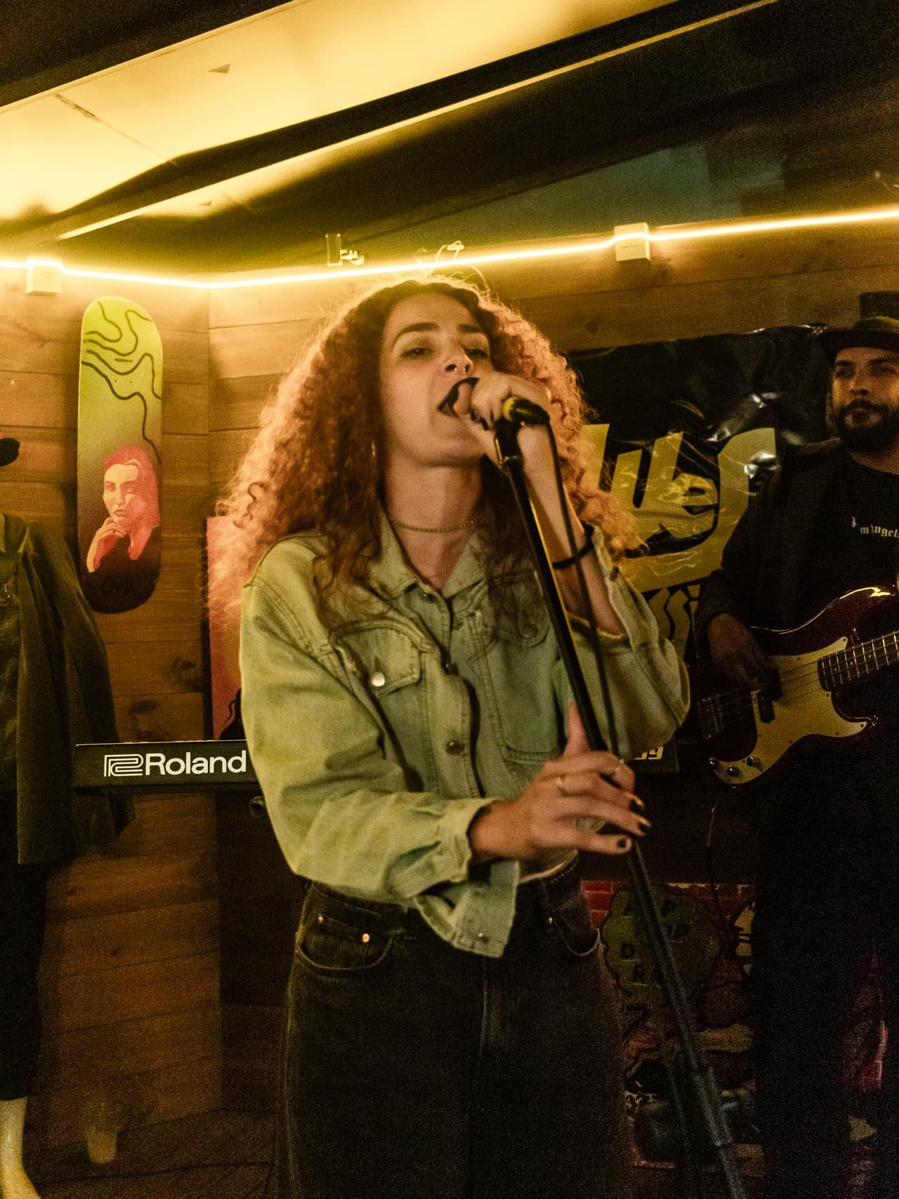Cantora de cachos e jaqueta jeans canta ao pedestal com baixista e banner Blues Session ao fundo