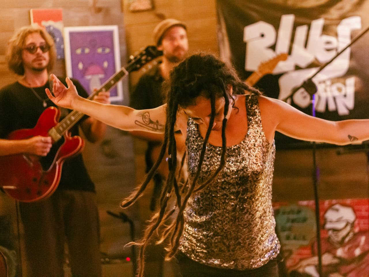 Cantora de dreads e blusa de paetês abre os braços no palco, com guitarristas e banner Blues Session atrás