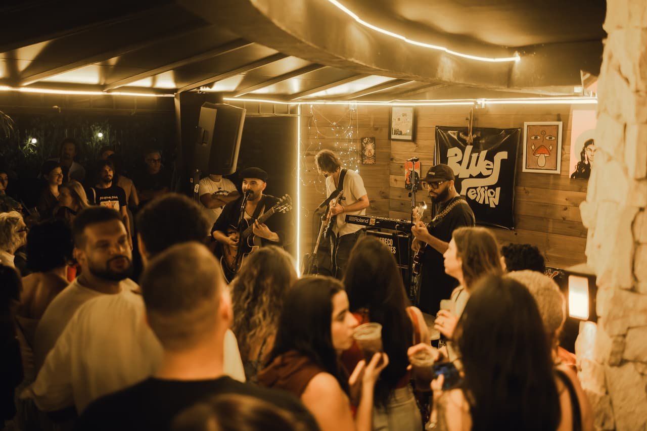 Vista do meio do público para o palco: salão cheio, luzes de LED no teto e banda tocando ao fundo