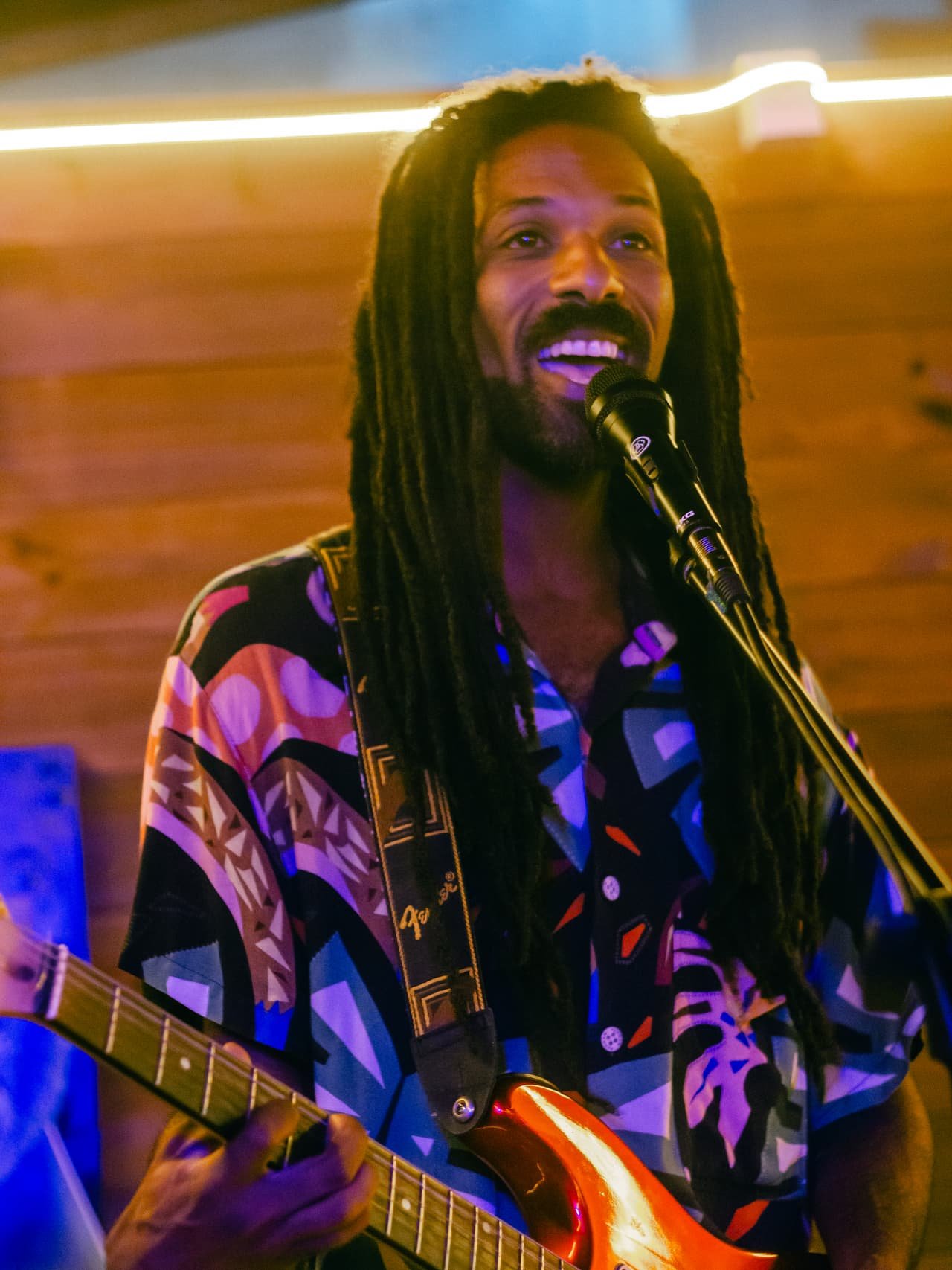 Cantor de dreads e camisa estampada sorri ao microfone enquanto toca uma guitarra vermelha