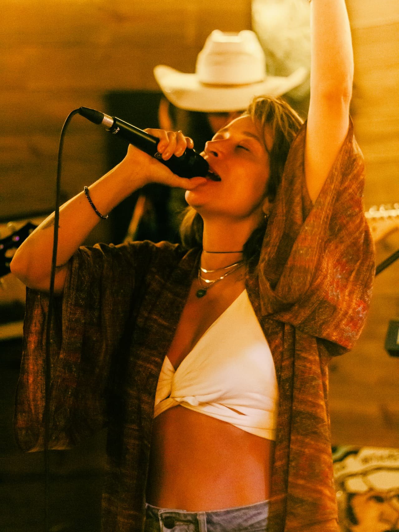 Cantora de chapéu de cowboy branco ergue o braço e canta colada ao microfone sob luz dourada