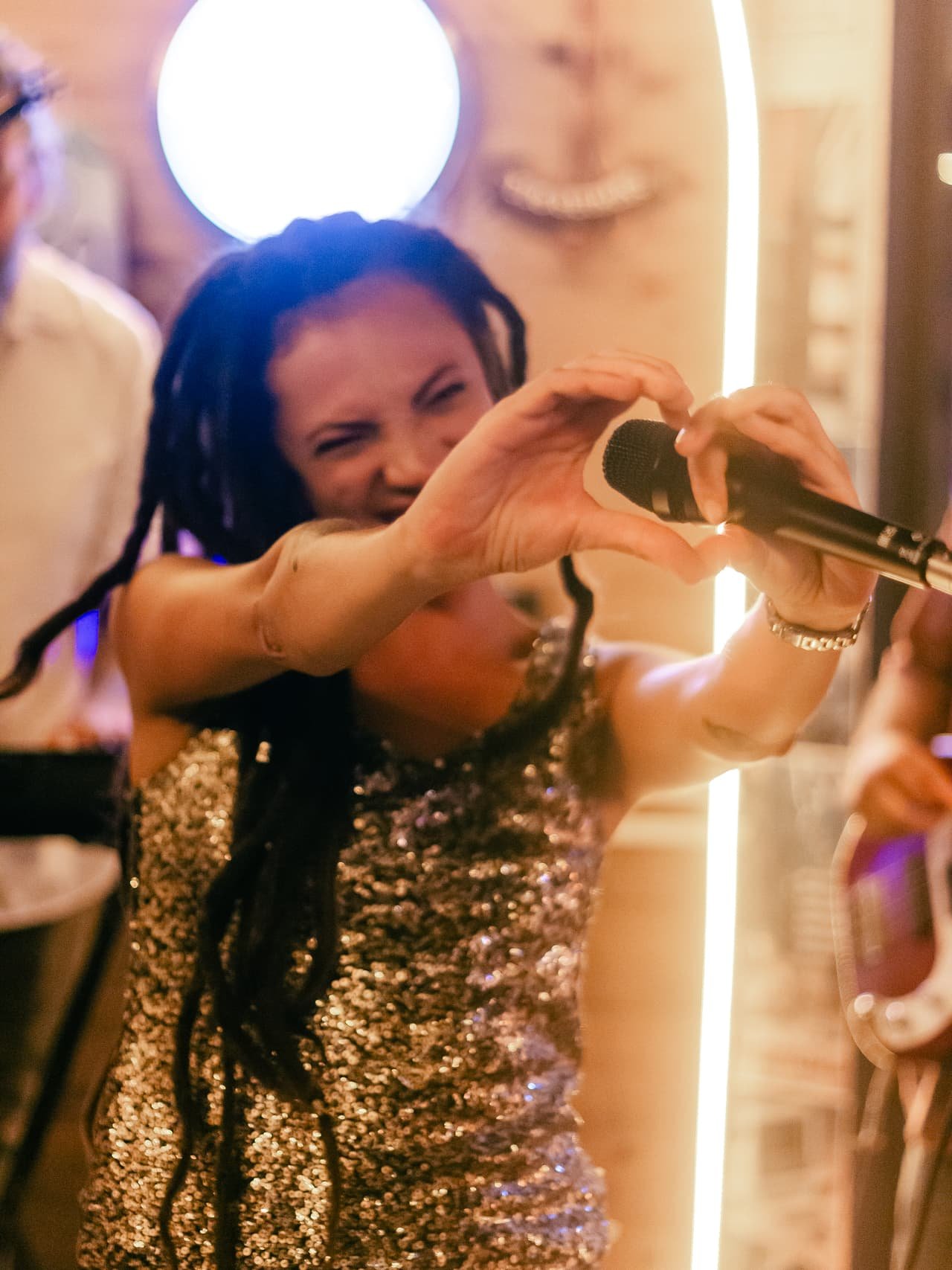 Cantora de dreads e blusa de paetês faz um coração com as mãos segurando o microfone