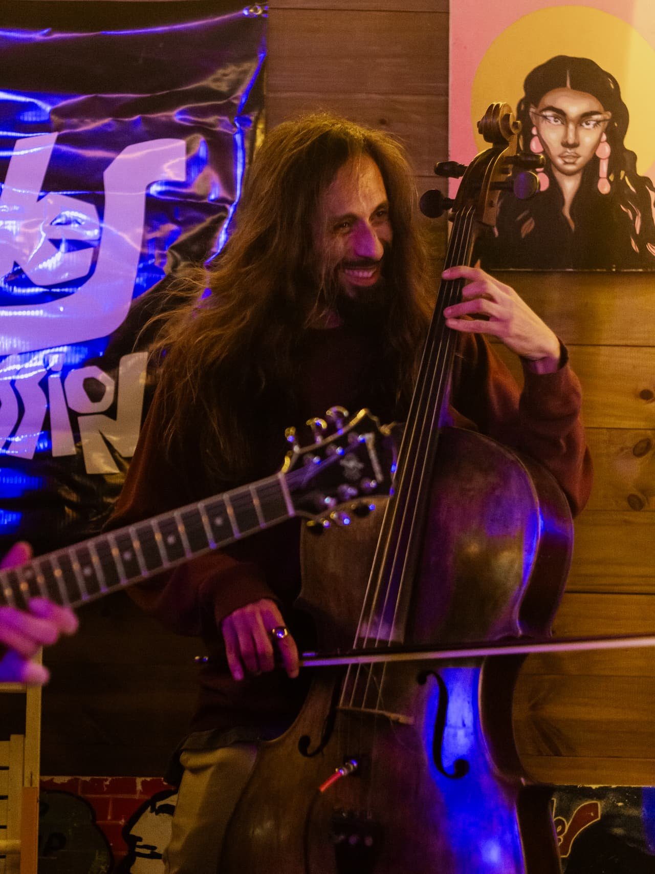 Músico de cabelos longos toca contrabaixo acústico sob luz azul e roxa, com uma guitarra em primeiro plano