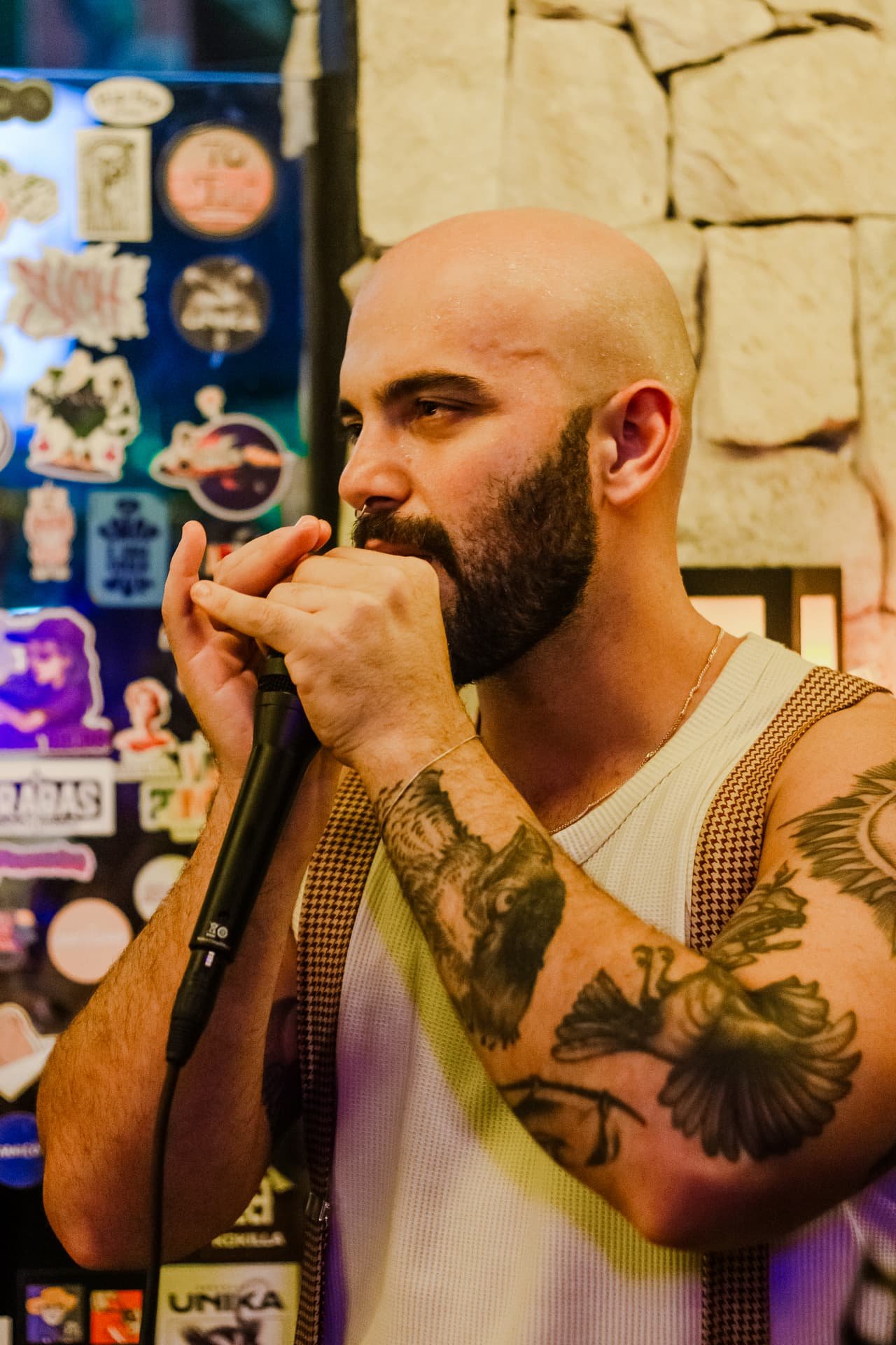 Gaitista careca e barbudo de braços tatuados toca gaita ao microfone diante de parede com adesivos
