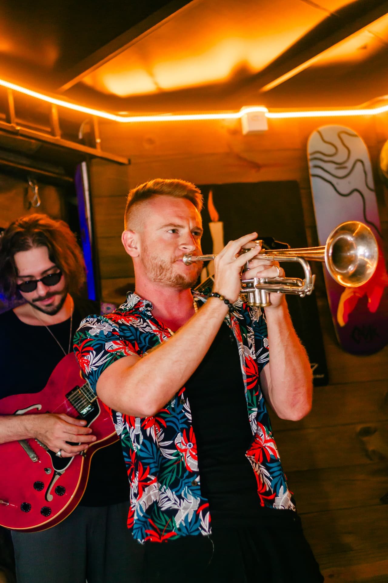 Trompetista de camisa florida toca de olhos fechados ao lado de um guitarrista, sob luz laranja
